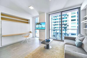 A modern living room with a large window, a flat-screen TV, a sofa, a coffee table, and built-in shelves. - Downtown Studio | Rooftop Pool | Walk to Bayside (Miami)
