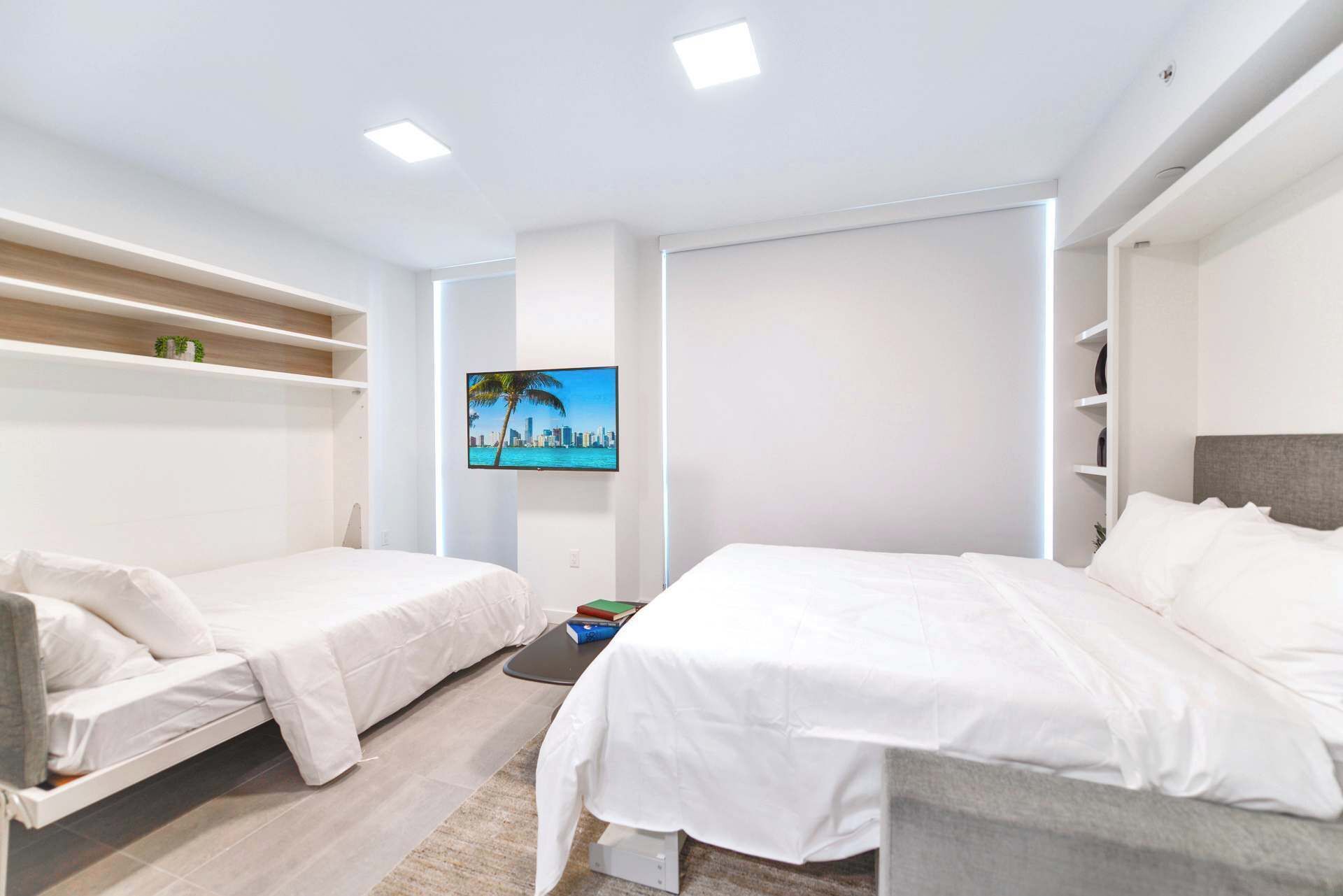 Moderni hotellihuone, jossa on kaksi sänkyä, taulutelevisio, sisäänrakennetut hyllyt ja näkymä kaupunkimaisemaan.