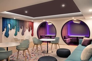Um lounge moderno com uma mesa redonda, cadeiras, um sofá e um elemento decorativo colorido na parede.