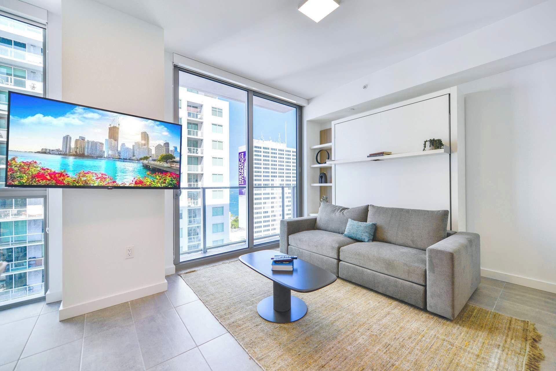 Ein modernes Wohnzimmer mit grauem Sofa, Couchtisch, Flachbildfernseher und einem großen Fenster mit Blick auf die Stadt.