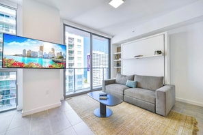 Ein modernes Wohnzimmer mit grauem Sofa, Couchtisch, Flachbildfernseher und einem großen Fenster mit Blick auf die Stadt.