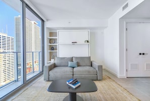 A modern living room with a grey sofa, a small coffee table with books and remotes, a white shelving unit, and a large window with a city view. - Downtown Studio | Rooftop Pool | Walk to Bayside (Miami)