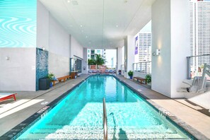 Une piscine à l’eau claire, entourée d’un bâtiment moderne doté de grandes fenêtres et offrant une vue sur d’autres immeubles.