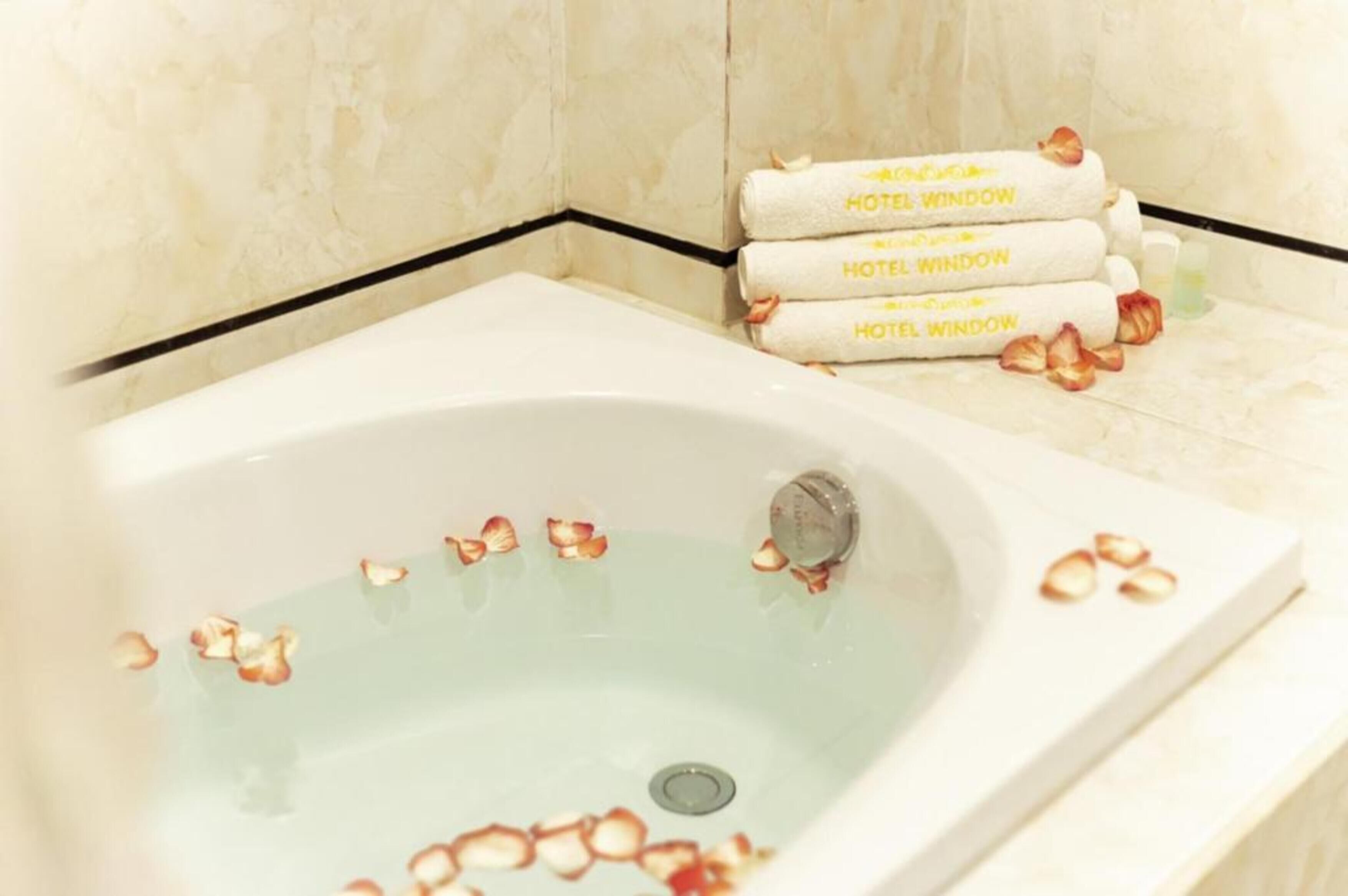 Eine Wanne mit darin treibenden Rosenblüten, ein Handtuchhalter mit Handtüchern, auf denen "HOTEL WINDOW" steht, und eine Wand mit Marmorfliesen.