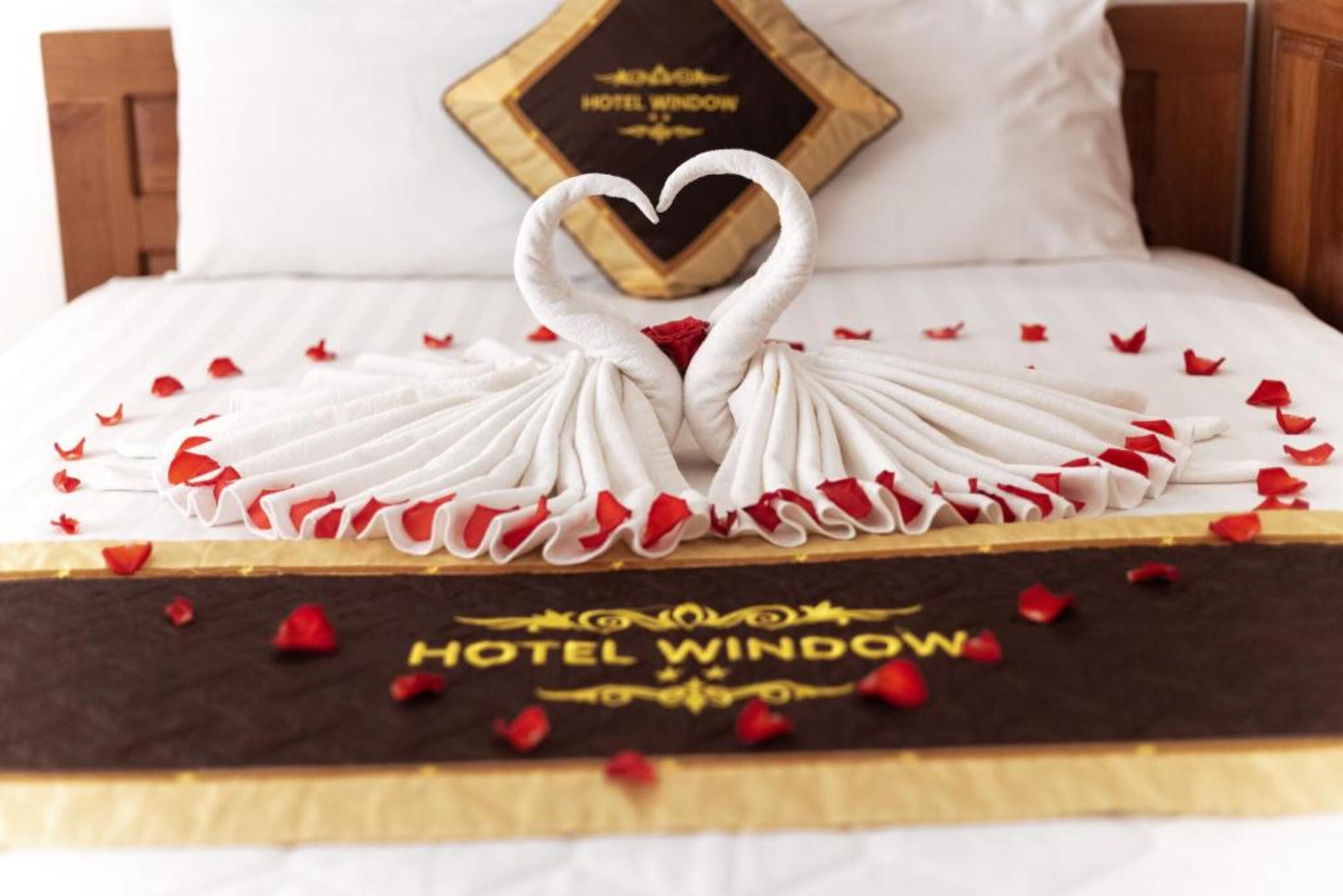 A bed with a heart-shaped swan decoration made of white fabric, surrounded by red rose petals, and a pillow with "HOTEL WINDOW" written on it.
