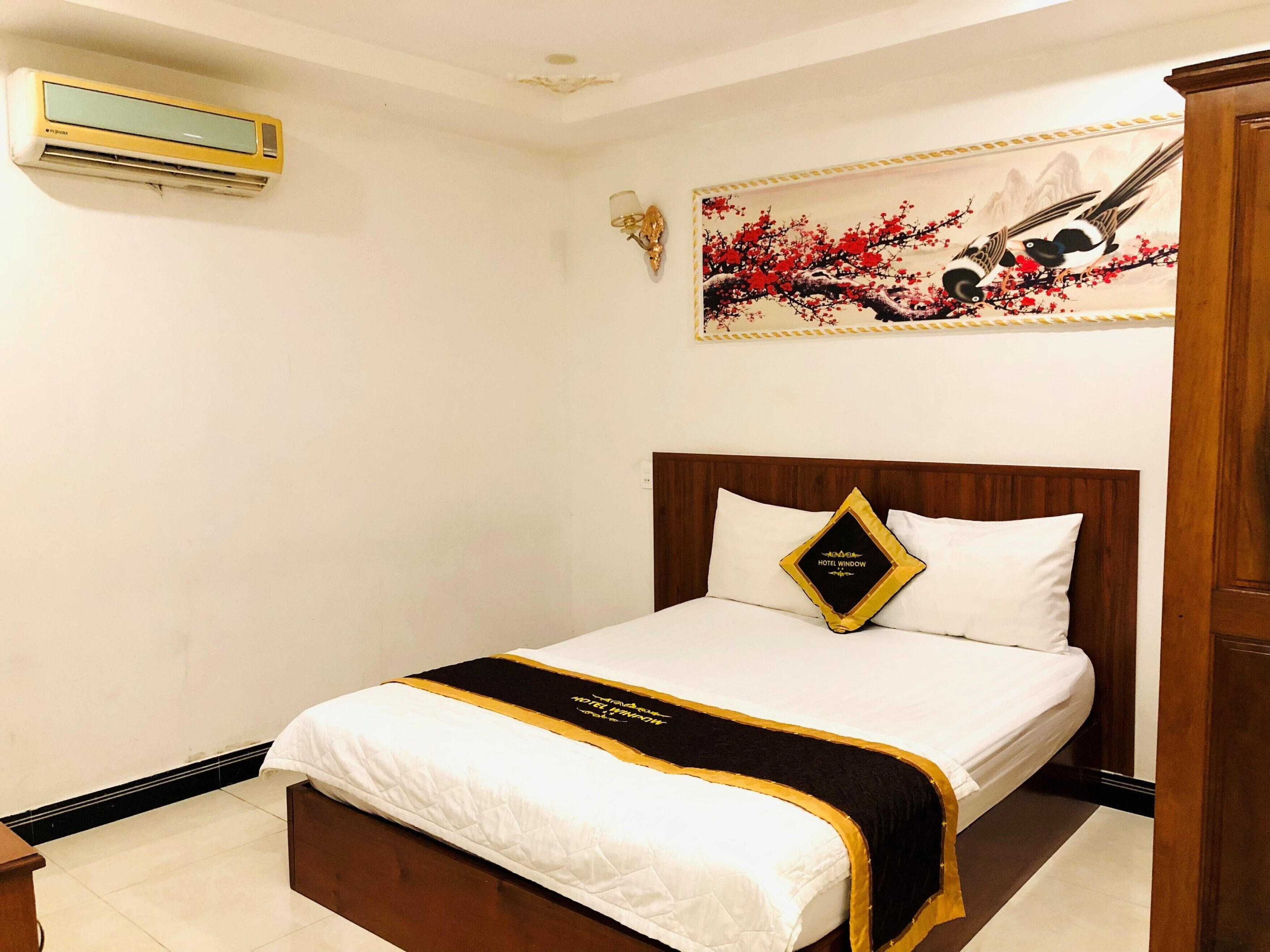 A hotel room with a bed, a wall-mounted air conditioner, a decorative painting, and a wooden headboard.