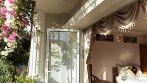 A room with a bed, a small table, and a chair. There is a window with curtains and a view of greenery outside. - Window 1 Hotel (Kon Tum)
