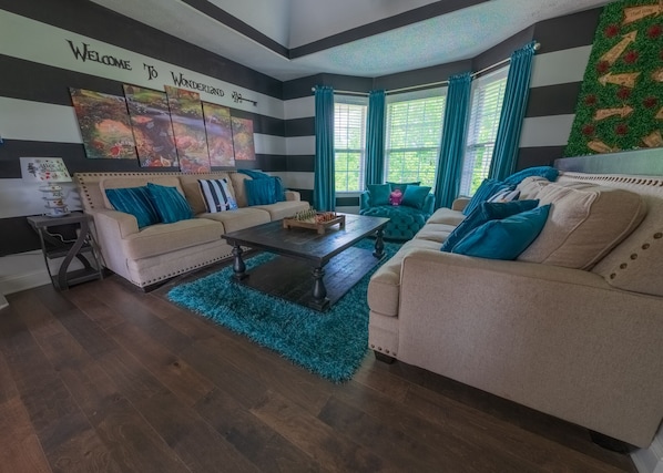 A living room with striped walls, a 'Welcome to Wonderland' sign, two sofas with teal cushions, a coffee table, and a window with curtains.
