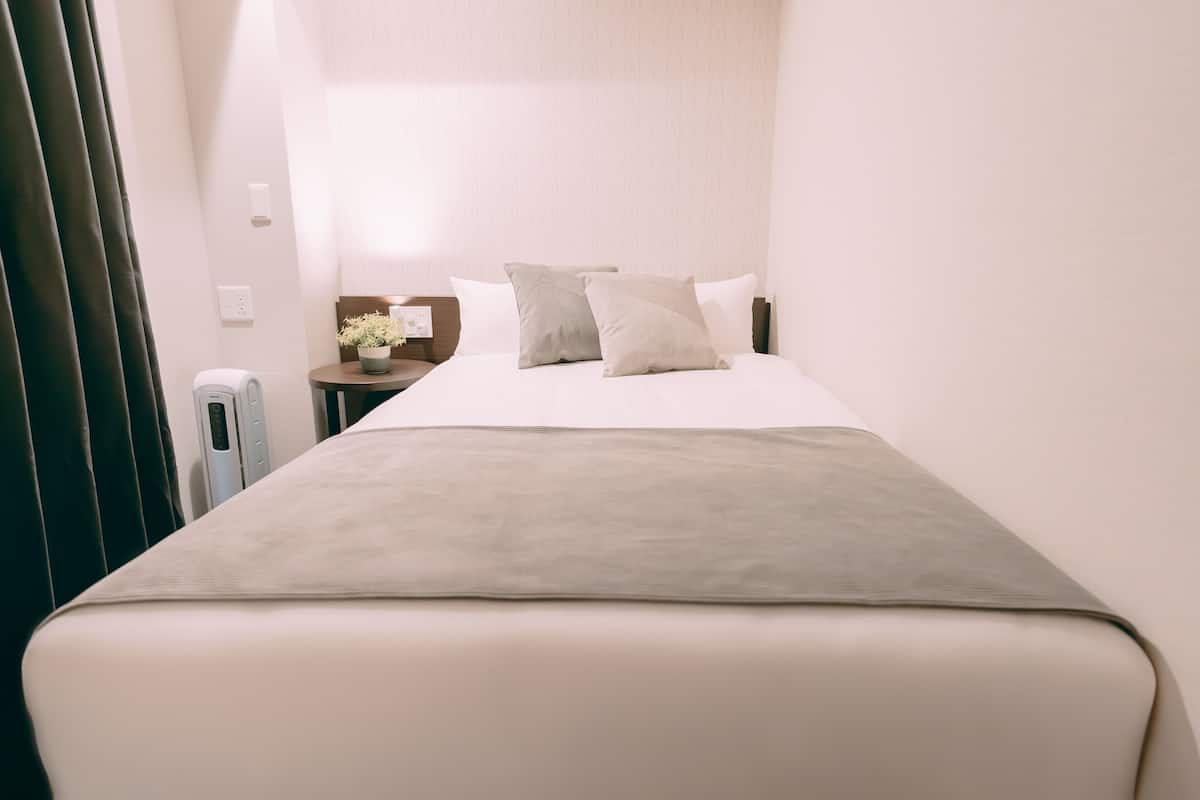 Una cama bien hecha con un edredón gris y dos almohadas, una mesita de noche con una lámpara y una pequeña planta, y una ventana con cortinas.