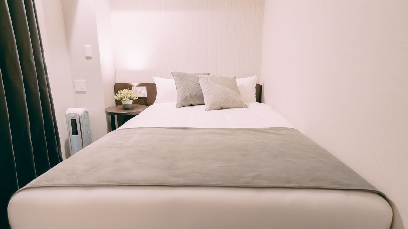 Una habitación de hotel con cama, mesita de noche, lámpara y una planta pequeña en maceta.