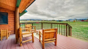 Una terraza de madera con mobiliario exterior, vista a un campo verde y montañas distantes.