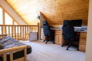 Ein gemütliches Schlafzimmer im Dachgeschoss mit Holzbalkendecke, einem Schreibtisch mit zwei schwarzen Bürostühlen, einem Computermonitor und einem Bett mit gemustertem Überwurf.