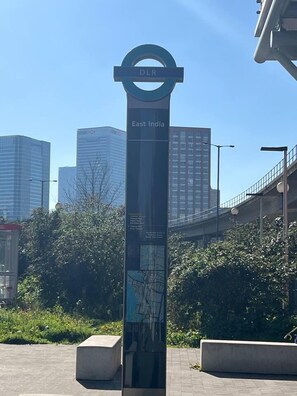 A tall, dark-colored sign with 'DLR East India' and a map on it, set against a cityscape with modern buildings and a clear sky. - Lovely & Cosy 2 Beds London Skyline View (London)