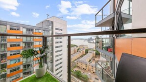 A high-rise apartment with a balcony, large windows, and a view of a river and other buildings. - Lovely & Cosy 2 Beds London Skyline View (London)