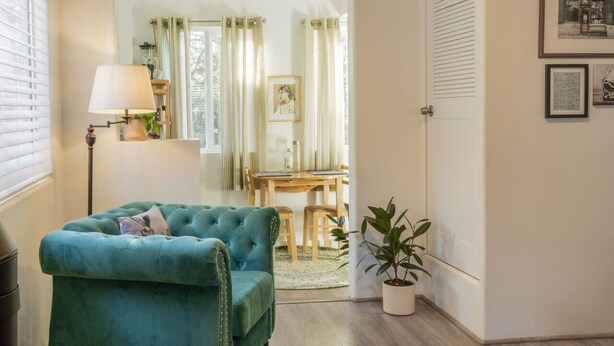 Et værelse med en teal farvet lænestol, en gulvlampe, et spisebord med stole, en potteplante og en dør, der fører til et andet rum.