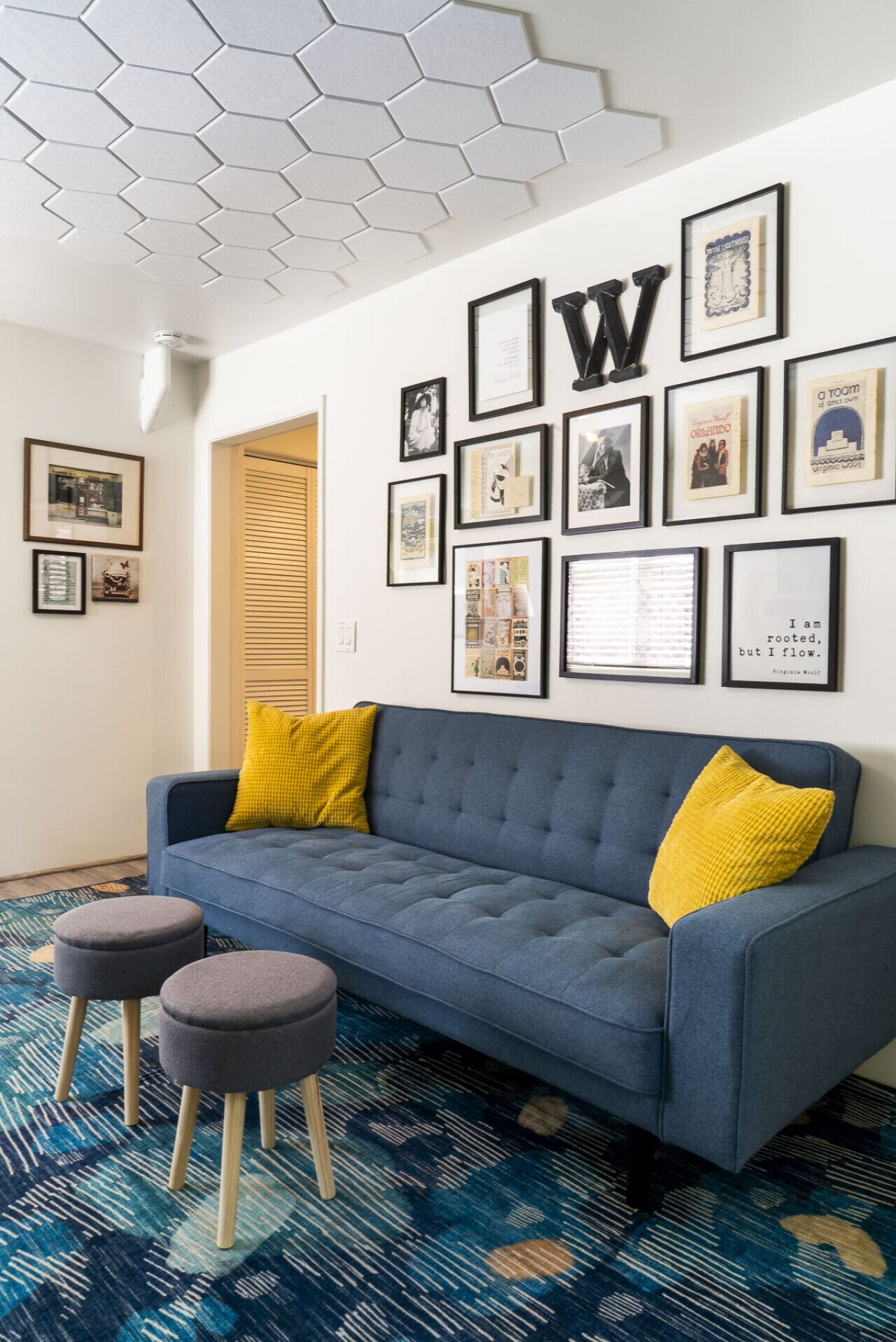 A modern living room with a blue sofa, two grey ottomans, and yellow cushions. The wall features a hexagonal ceiling and multiple framed pictures.