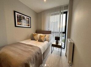 Quarto de hotel moderno com uma cama grande, uma mesinha de cabeceira, um quadro na parede e uma janela com persianas.