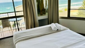 A hotel room with a large bed, a balcony with a view of the ocean, and a dining area with a table and chairs. - One Bed Room Ocean View With Surfers Holiday Accommodation - Aloha Apartments (Surfers Paradise)