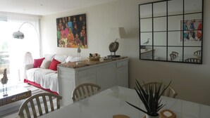 Une pièce bien éclairée, avec une table à manger, des chaises, un canapé blanc, un tableau et un miroir au mur.