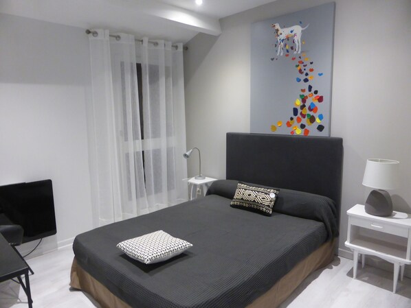 A modern bedroom with a large bed, a bedside table with a lamp, a chair, and a window with sheer curtains.