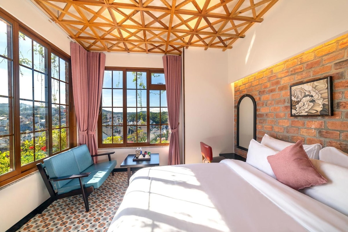 Habitación de hotel con una cama grande, un sofá azul, una mesita y vistas a un paisaje urbano a través de amplias ventanas.