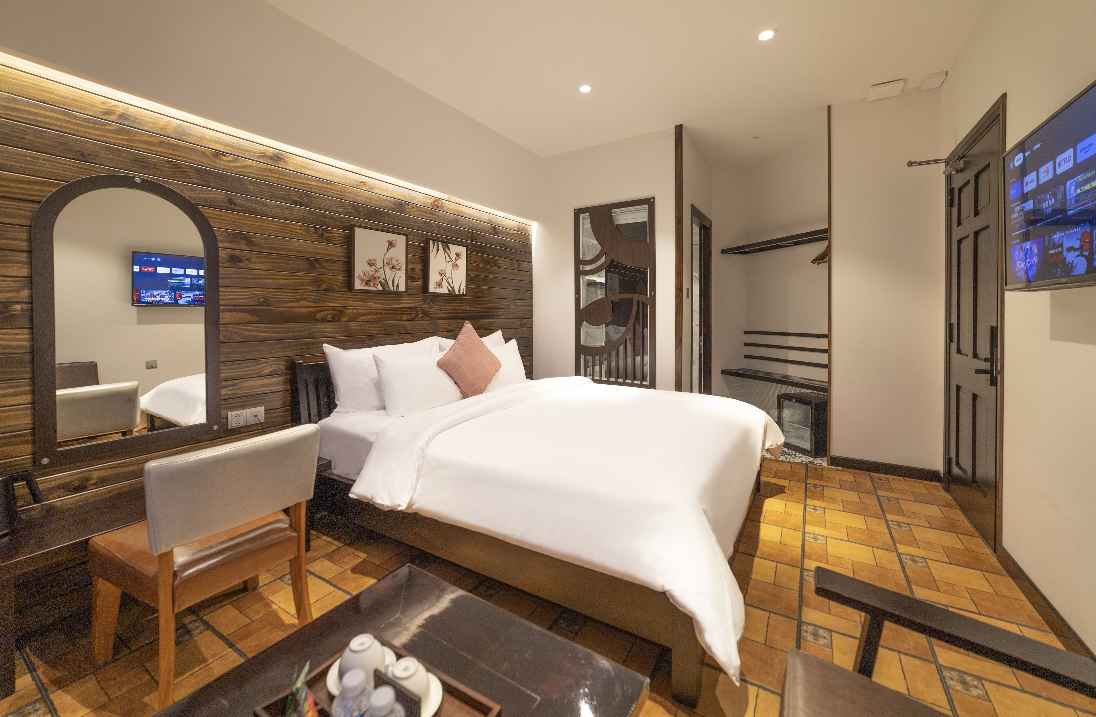 A modern hotel room with a large bed, a wooden headboard, a mirror, a television, and a small seating area.