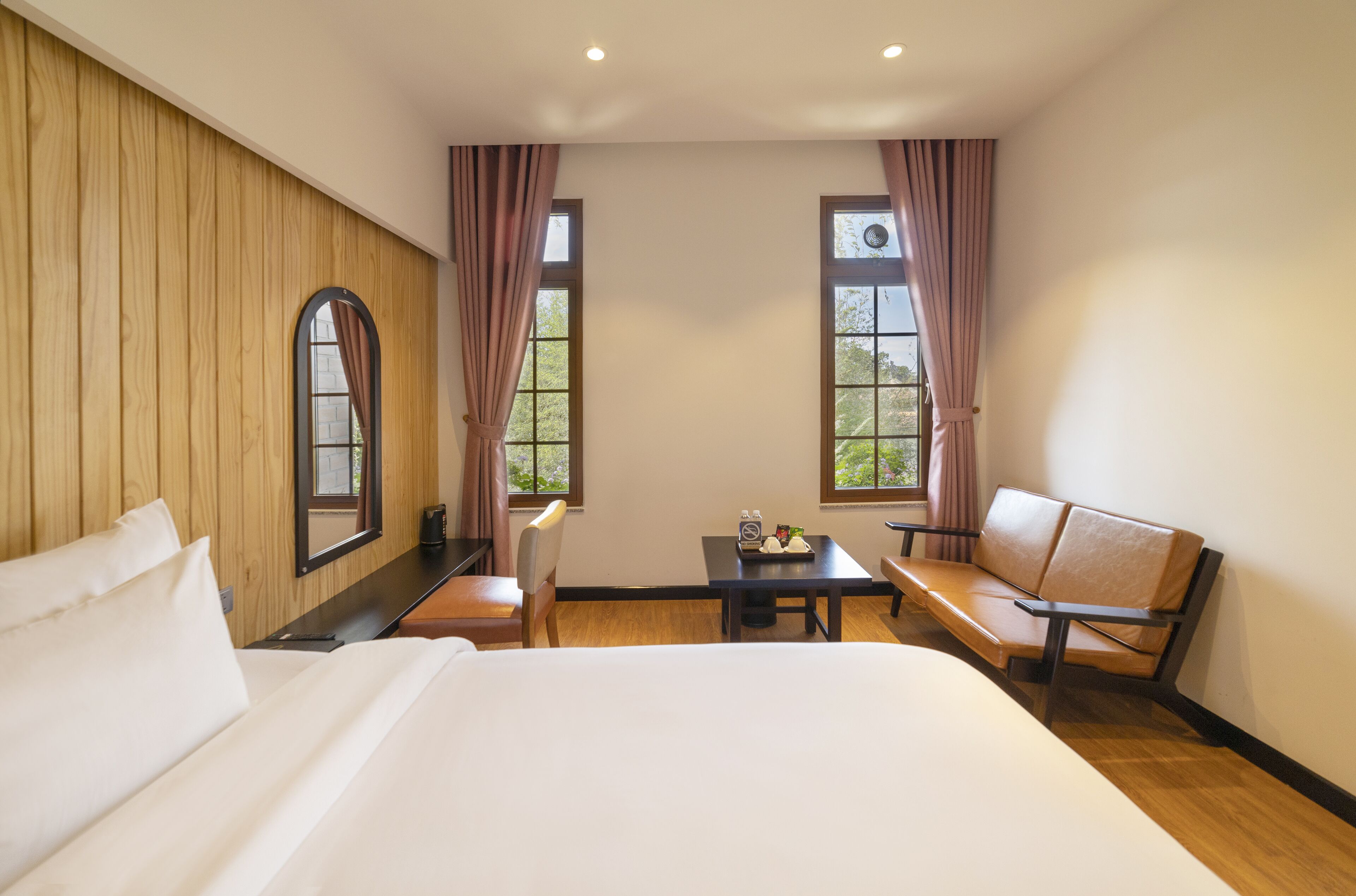 Quarto de hotel com uma cama grande, duas cadeiras, uma mesinha e duas janelas com cortinas.