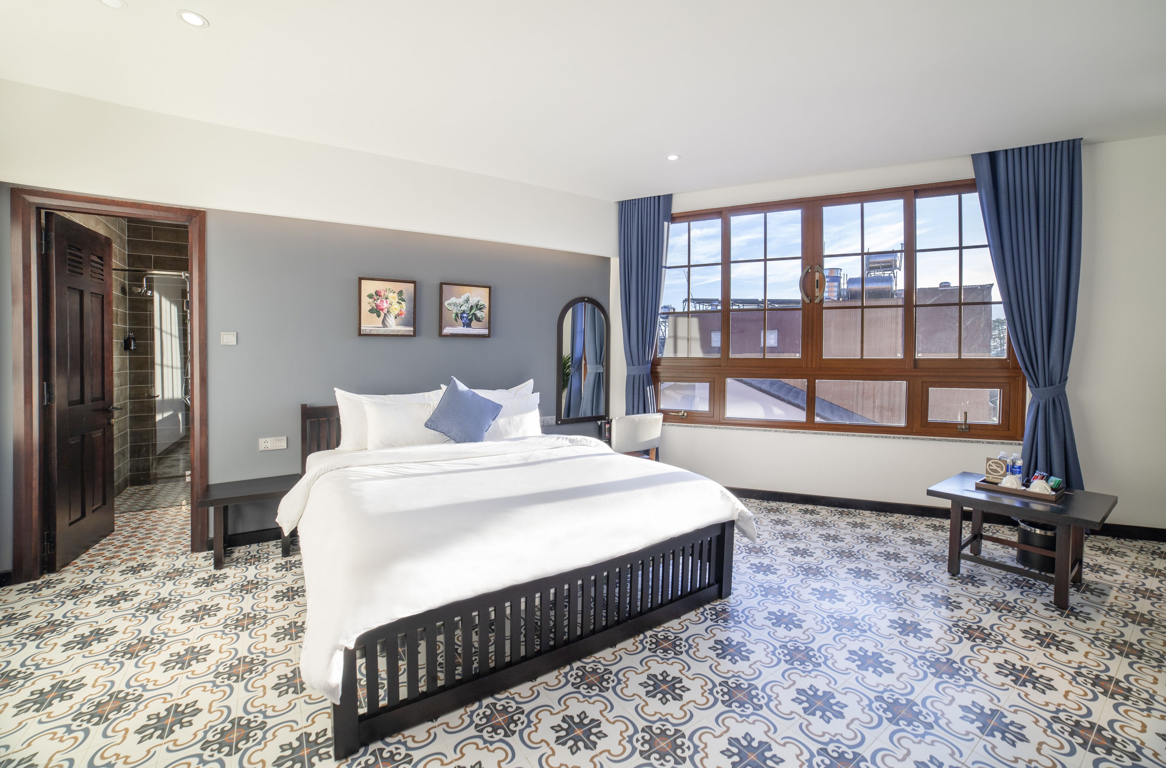 A bedroom with a large bed, a window with curtains, a small table, and a door leading to a bathroom.