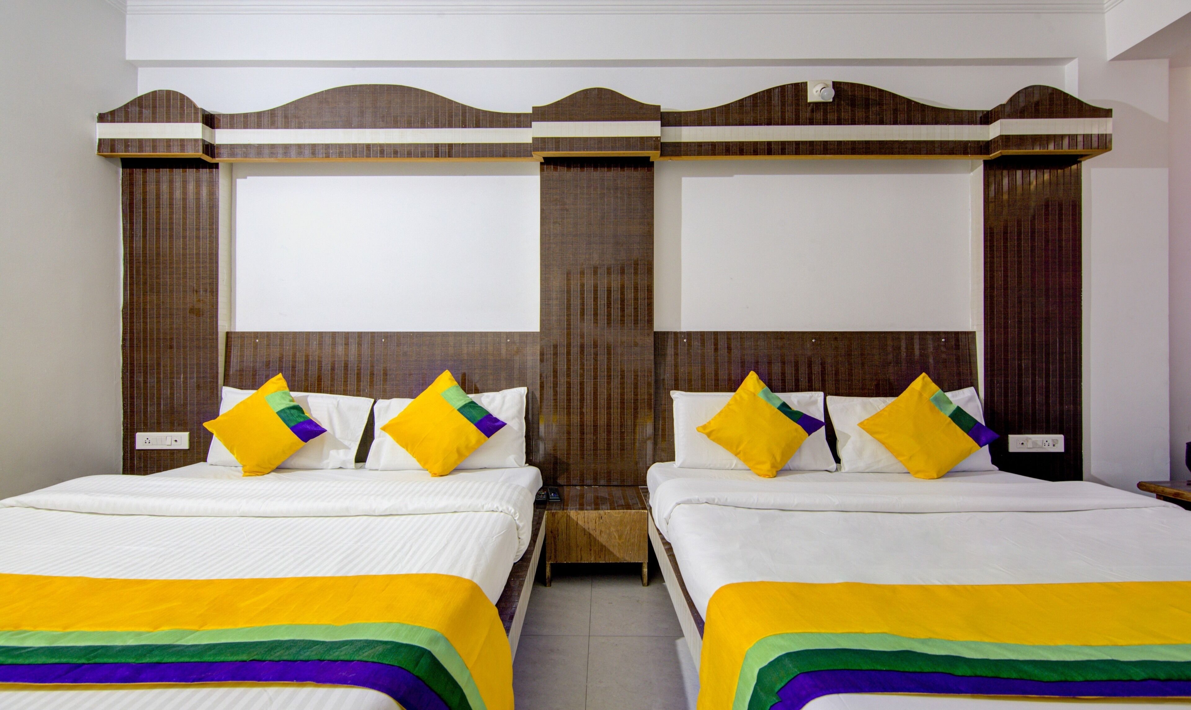 A hotel room with two beds, each with a white mattress and a colorful bedspread. The room has a wooden headboard with a decorative design.