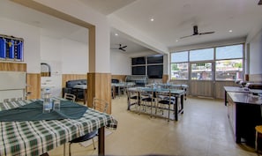 A spacious dining area with multiple tables and chairs, a kitchen area with a grill, and large windows providing a view of the outside. - Itsy Hotels Satya Shree (Reasi)