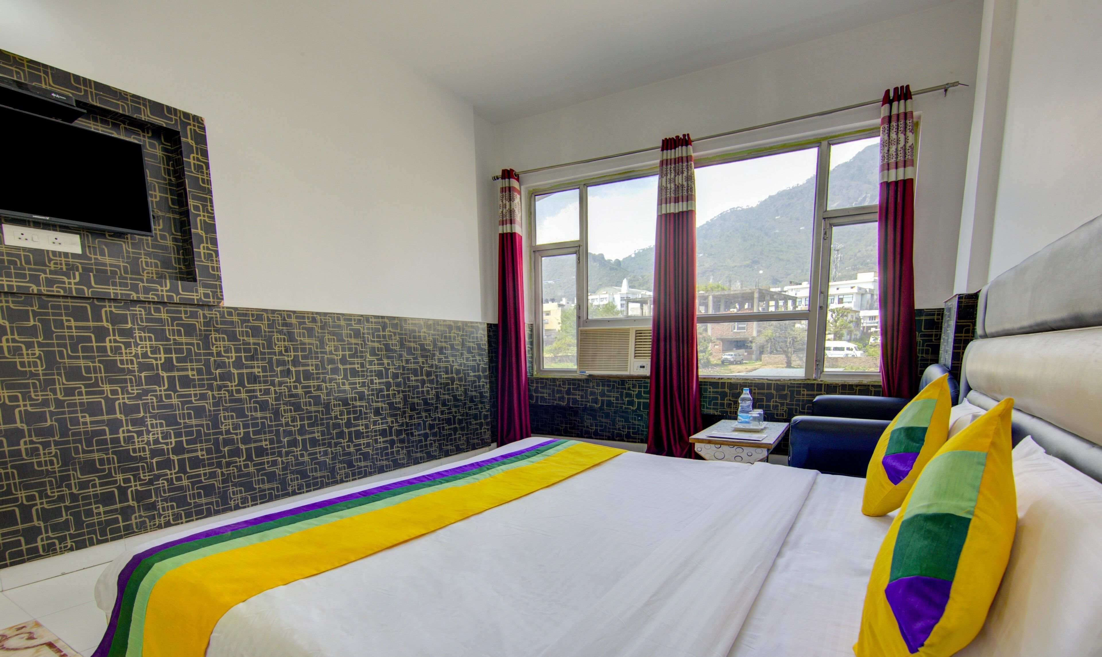 Quarto de hotel com cama, janela com cortinas, vista para prédios e televisão montada na parede.