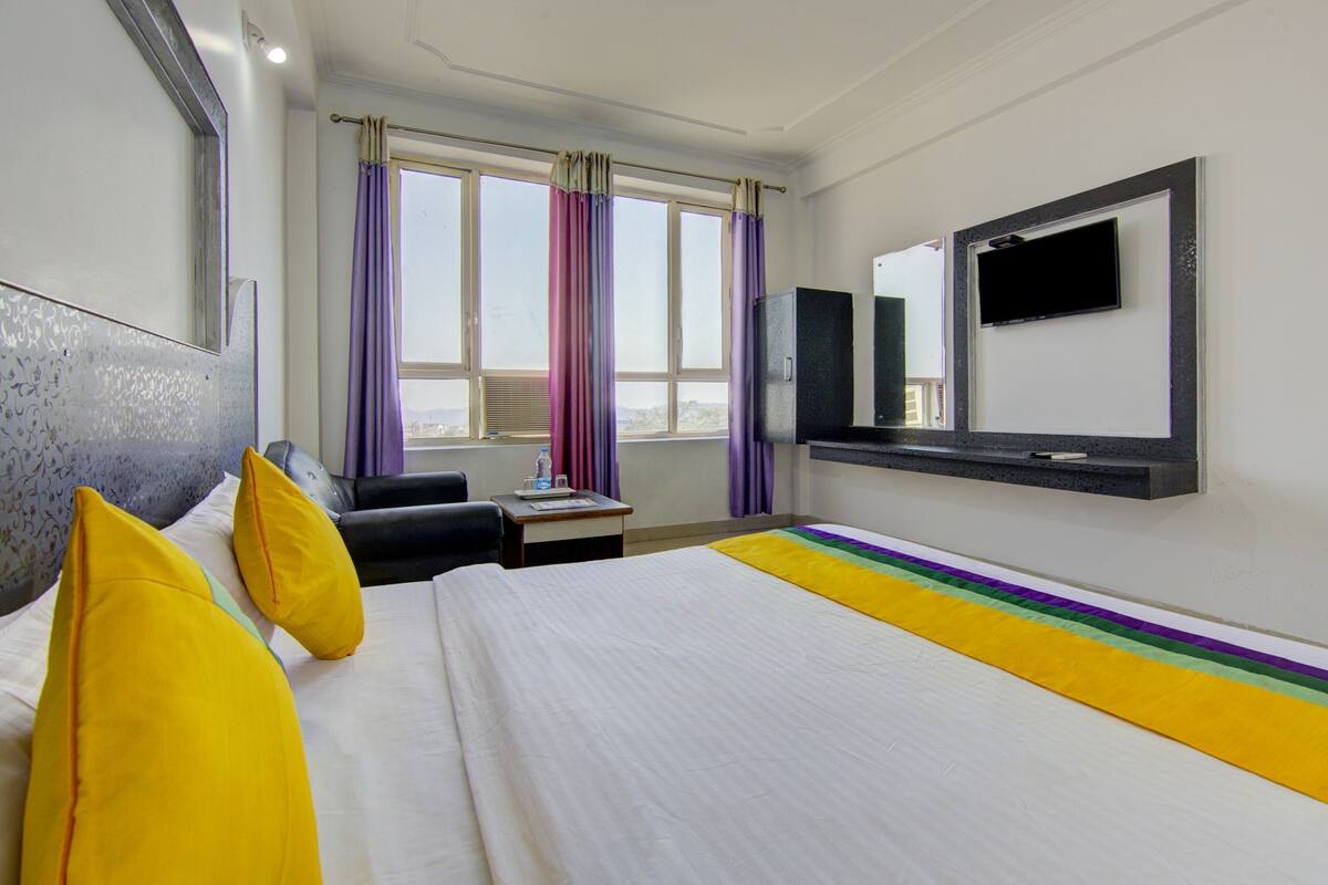 Habitación de hotel con cama, escritorio, silla, televisor y ventana con cortinas.