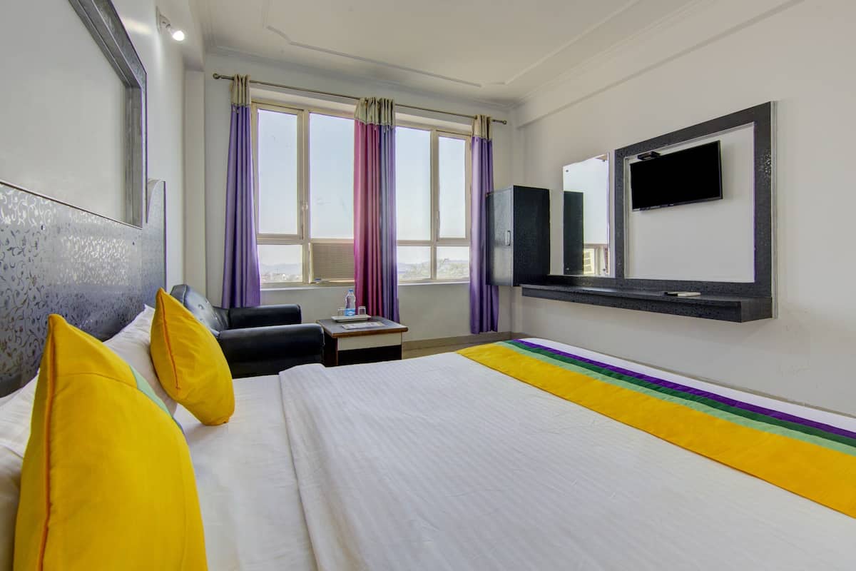 Quarto de hotel com cama, escrivaninha, cadeira, televisão e janela com cortinas.