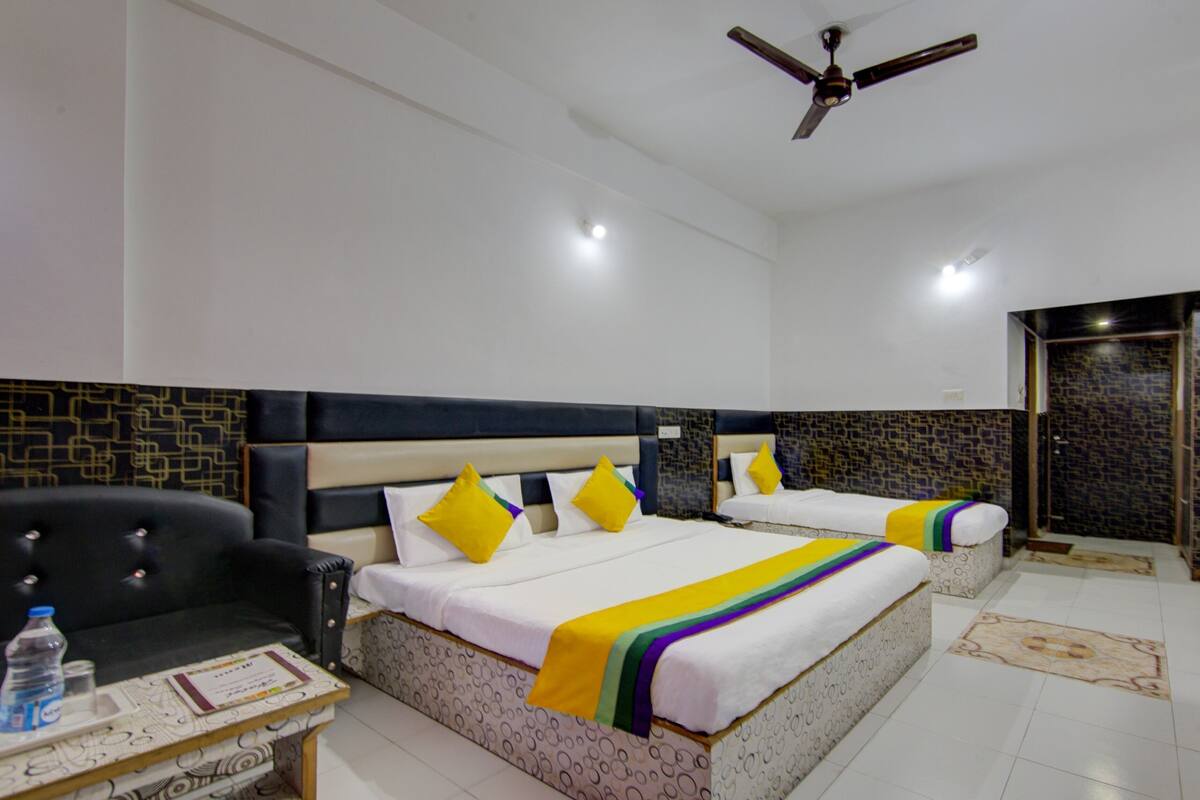 Una habitación de hotel con dos camas, un ventilador de techo, un sillón de cuero negro, una mesita con una botella de agua y una alfombra estampada.