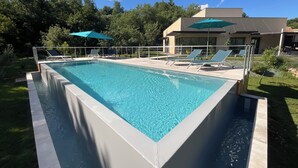 A rectangular swimming pool with a white border, surrounded by a metal fence. There are lounge chairs and a blue umbrella near the pool.