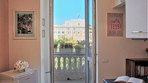 Ein Zimmer mit Blick auf ein Gebäude, ein gerahmtes Bild, ein kleiner Tisch mit einer Vase und ein Bücherregal mit Büchern.