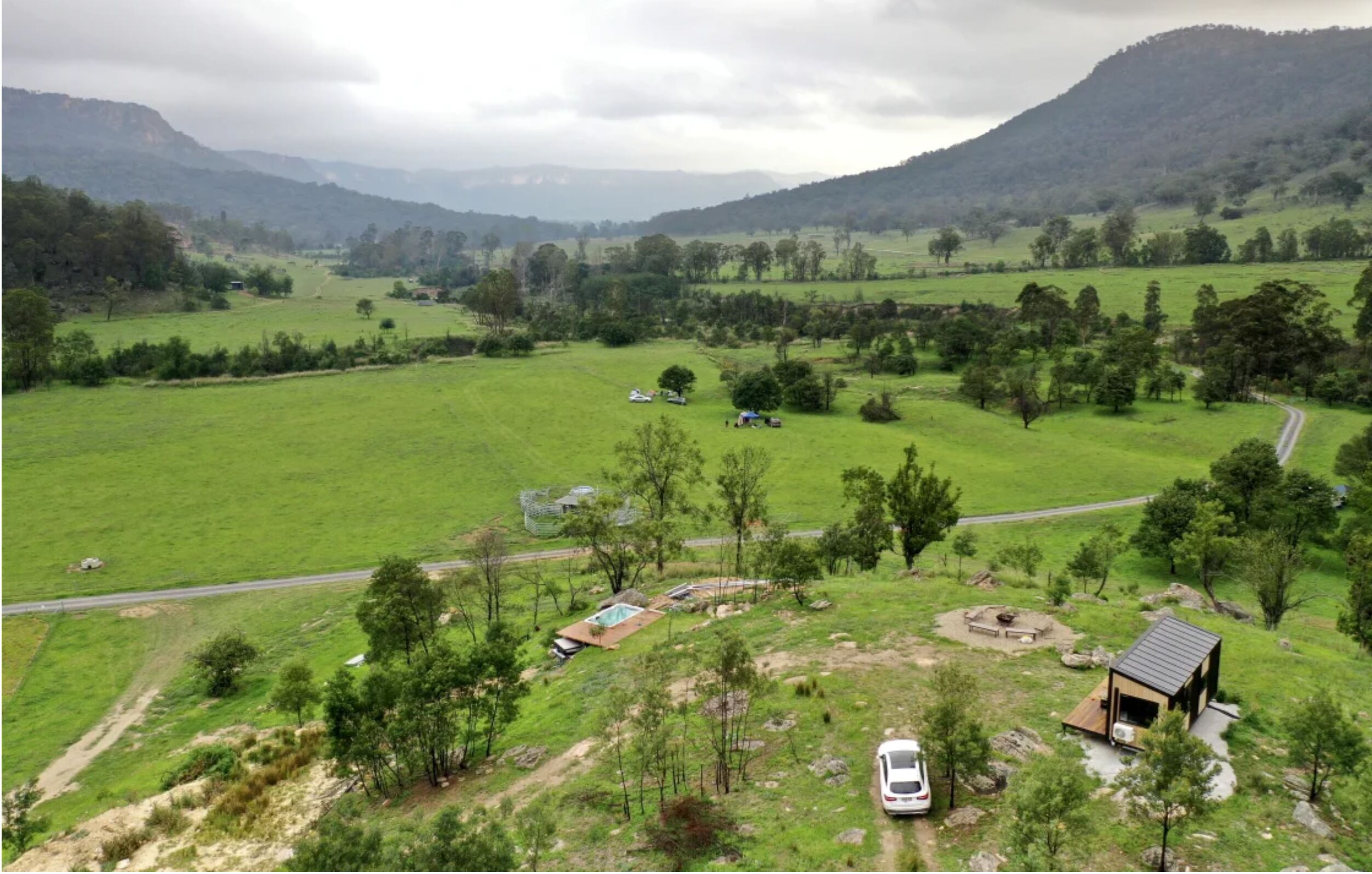 A rural landscape with a small cabin, a swimming pool, and a car parked in a green field.