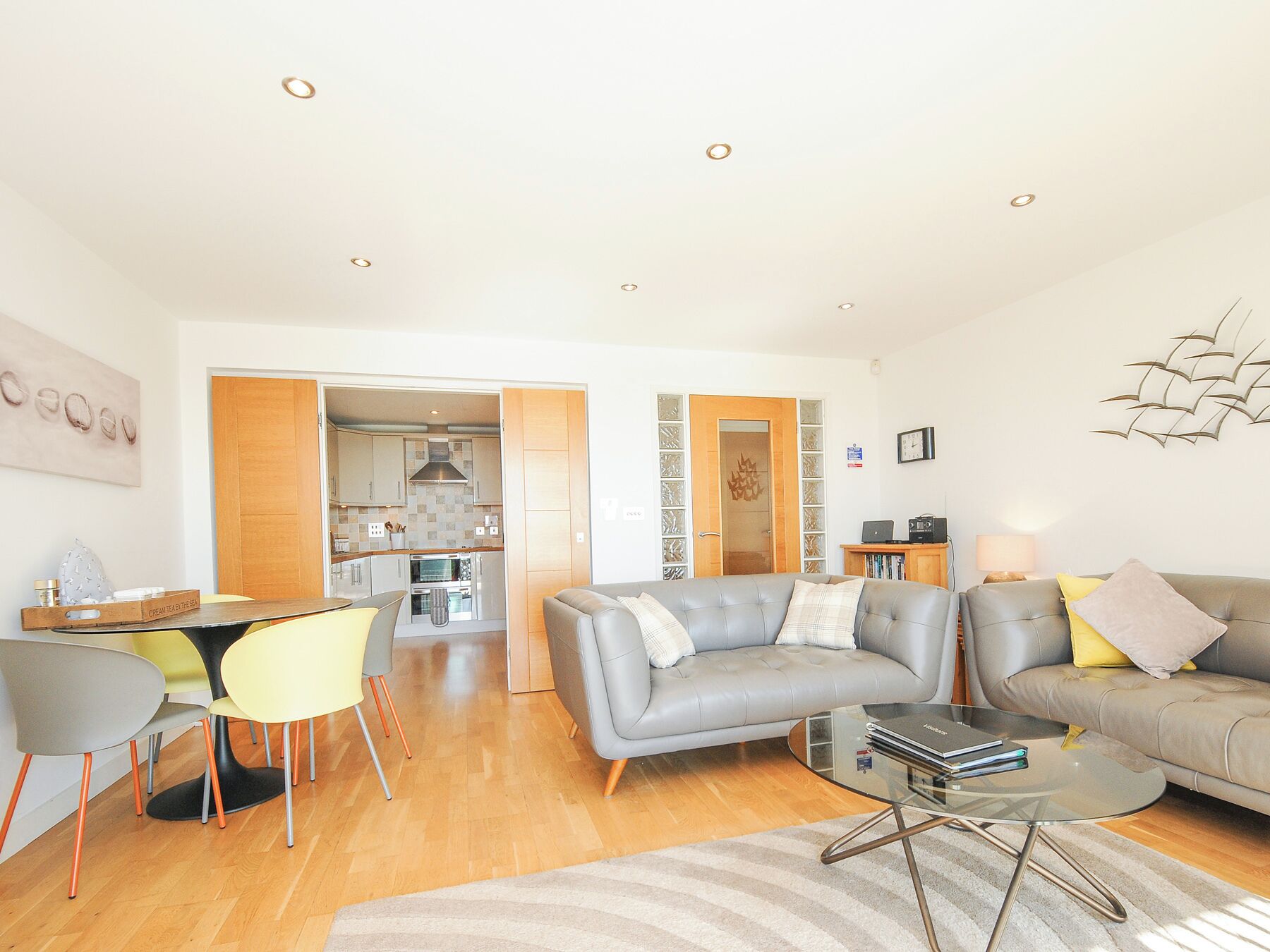 A modern living room with a grey sofa, a glass coffee table, and a dining area with a yellow chair.