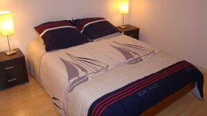 Un lit bien fait, recouvert d’une couvre-lit à motifs de voiliers, avec deux oreillers assortis, deux lampes de chevet et une petite table de nuit avec une lampe de chaque côté.