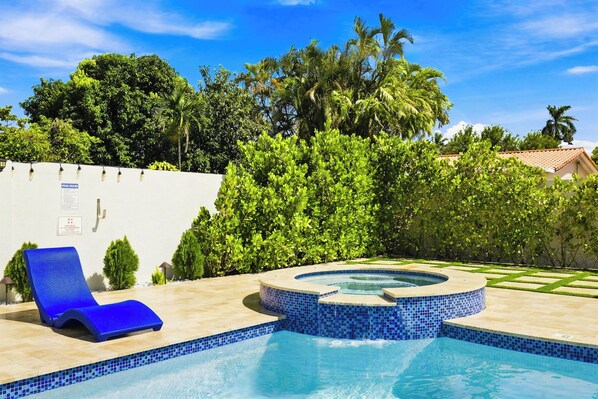 A swimming pool with a hot tub, surrounded by lush greenery and a clear sky.