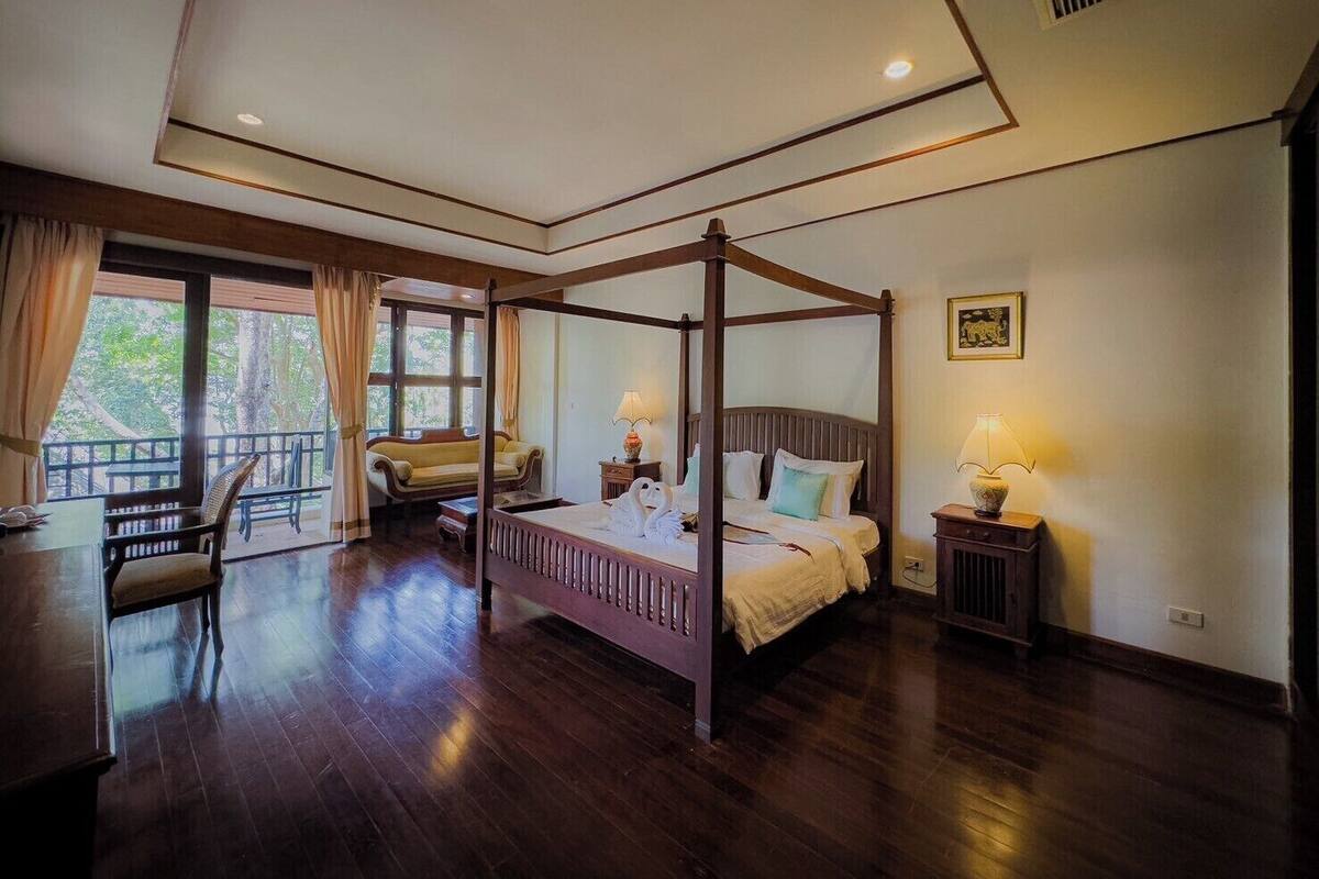 A four-poster bed with a wooden frame, a bedside table with a lamp, and a chair in a room with wooden flooring and large windows.