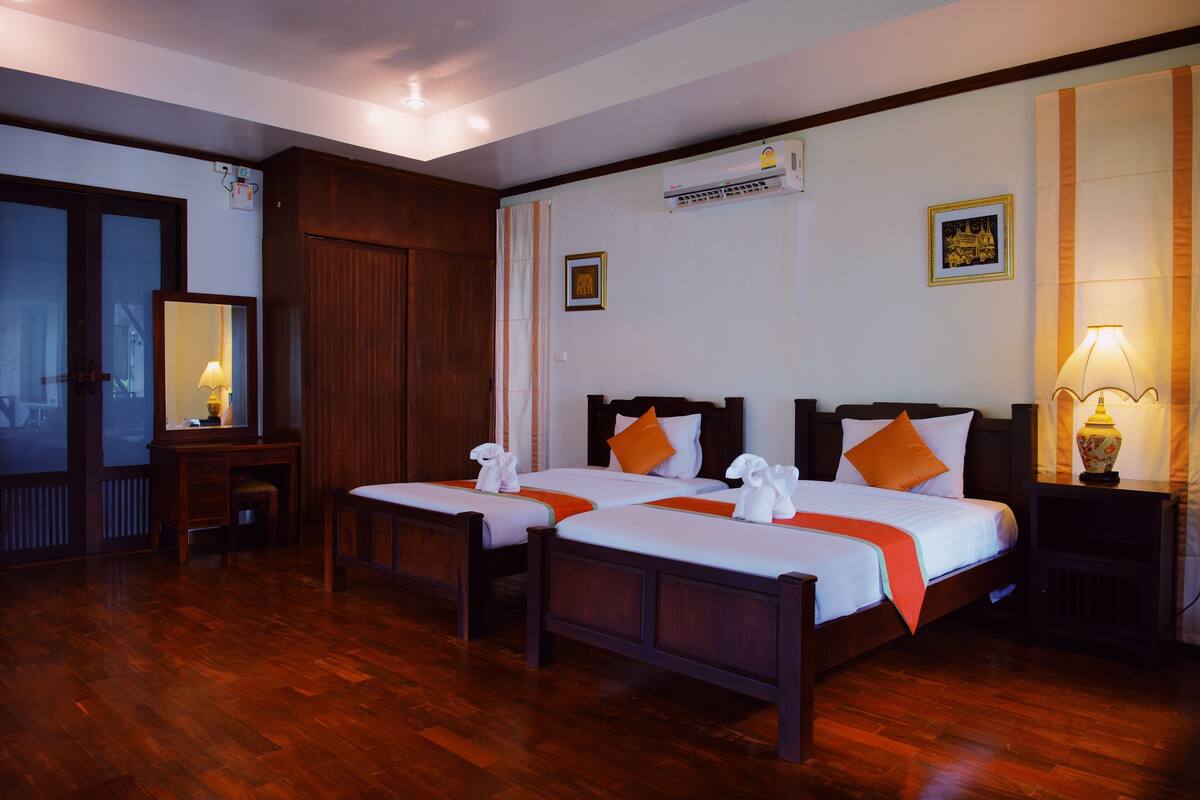 A hotel room with two beds, wooden flooring, a desk, a chair, a mirror, an air conditioner, and a nightstand with a lamp.