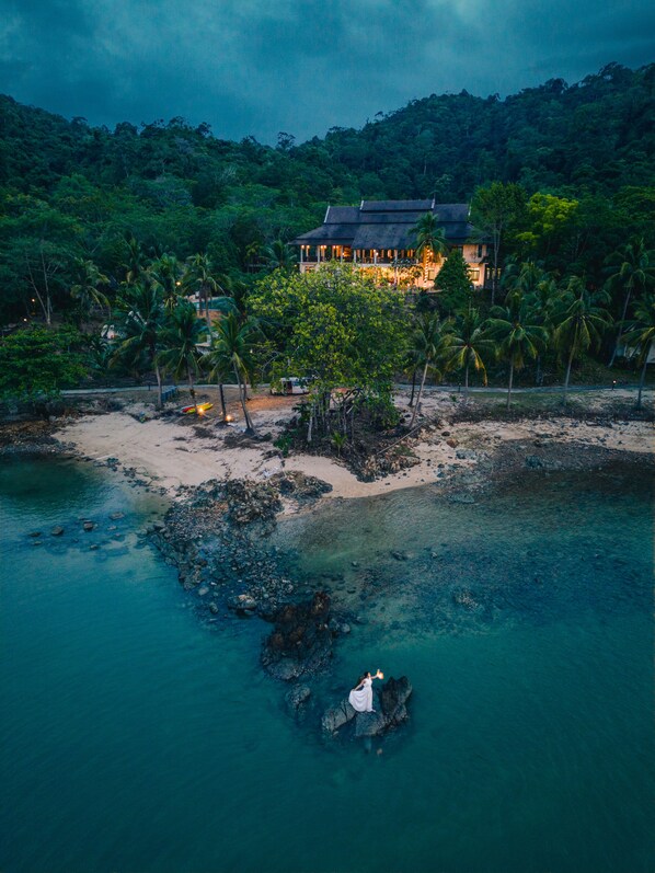 A coastal property with a beach, palm trees, and a house with a dark roof.