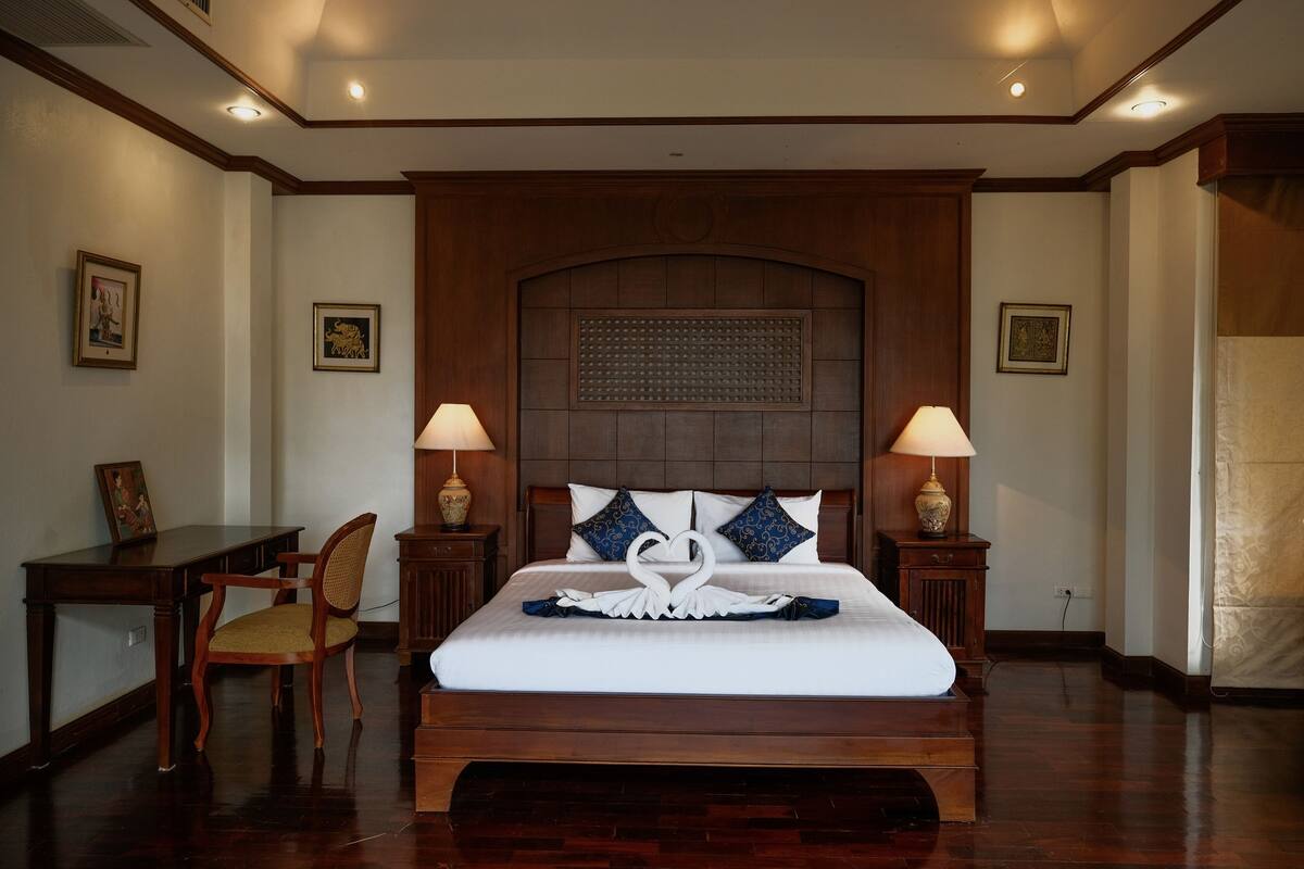 A spacious hotel room with a large bed, wooden headboard, two bedside lamps, a wooden desk, and a chair.