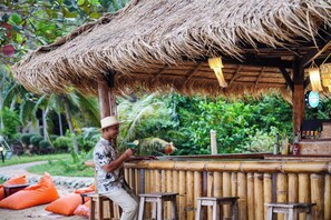 Zona de bar al aire libre con techo de paja, asientos de bambú y barra.