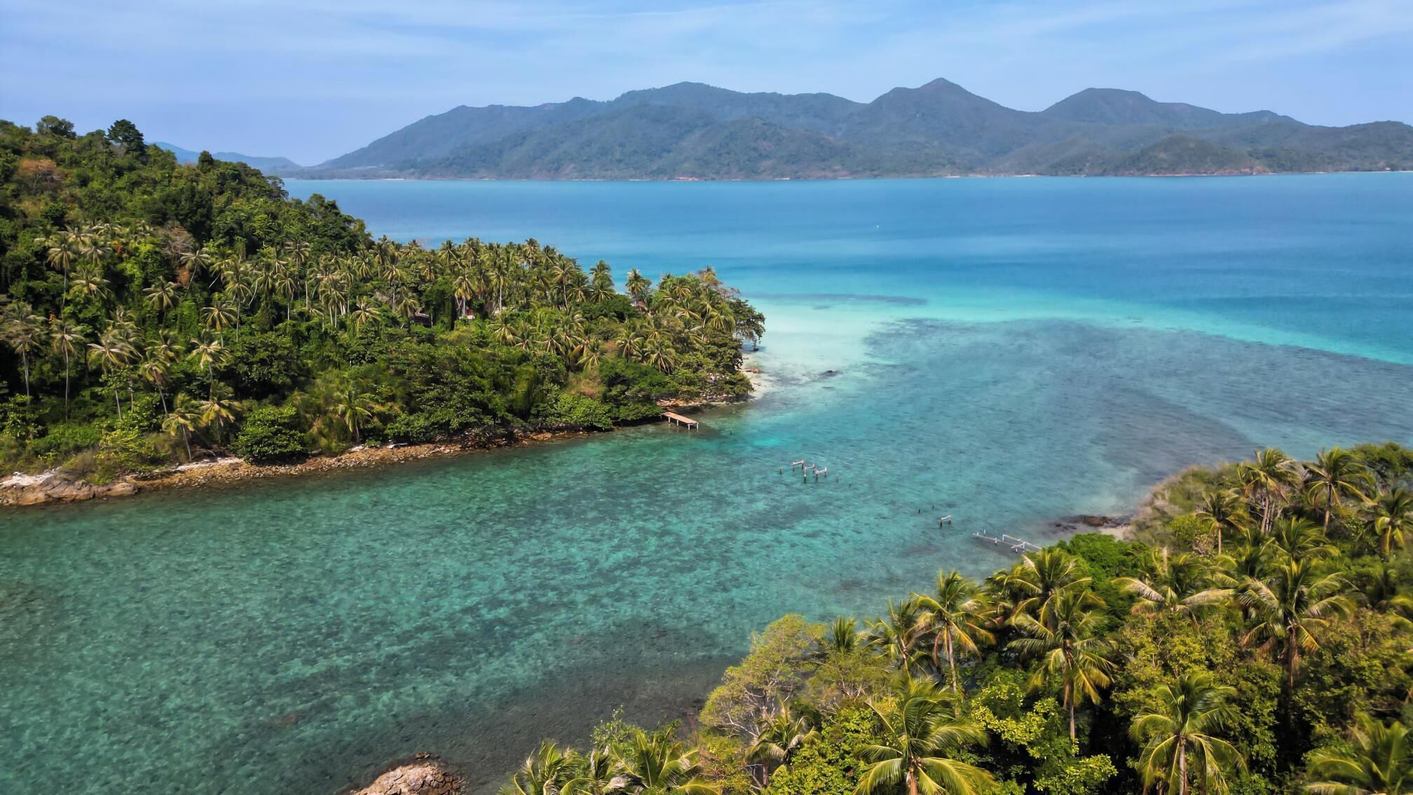 A tropical island with clear turquoise waters, lush greenery, and distant mountains.