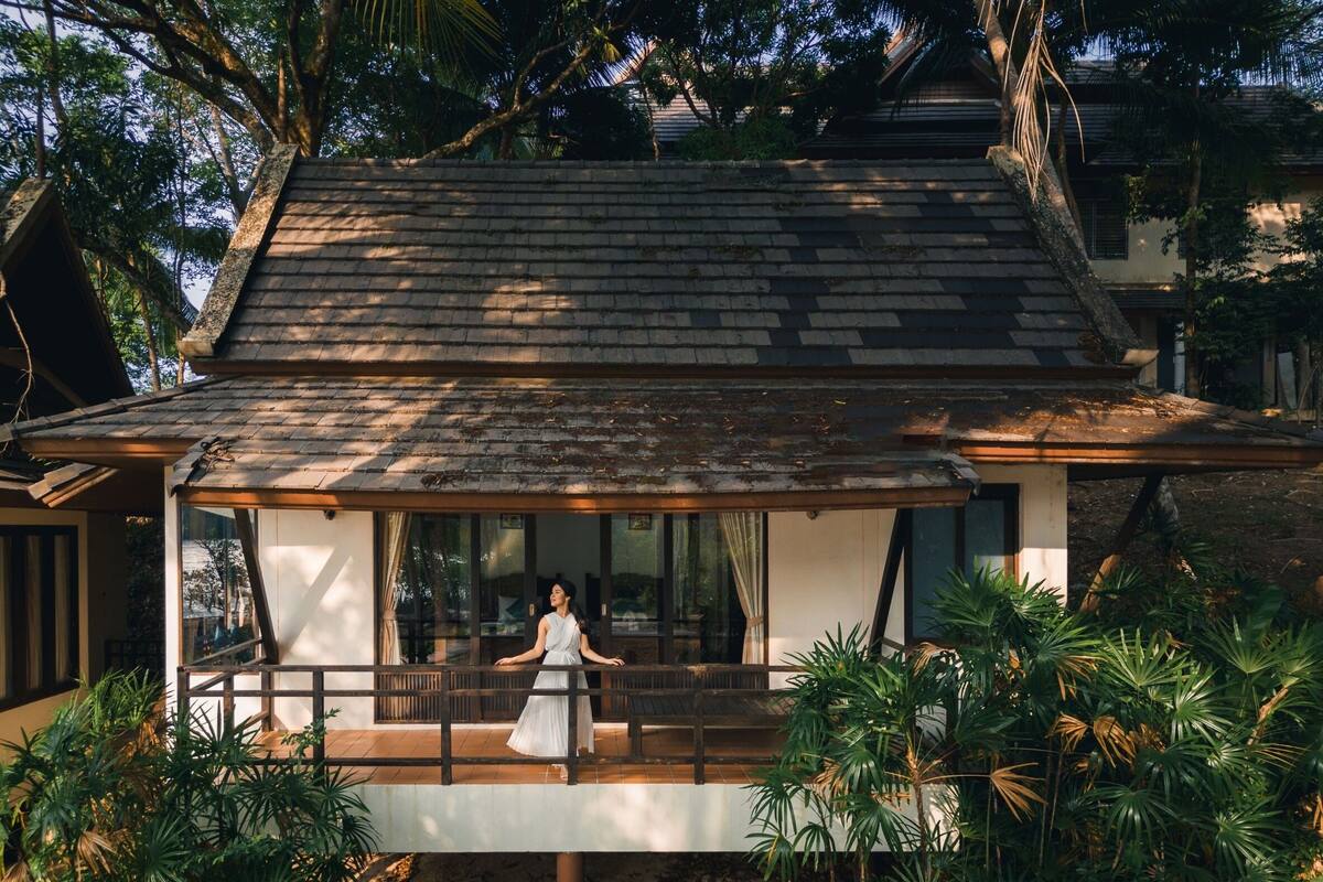 A traditional house with a wooden balcony and large windows, surrounded by lush greenery.