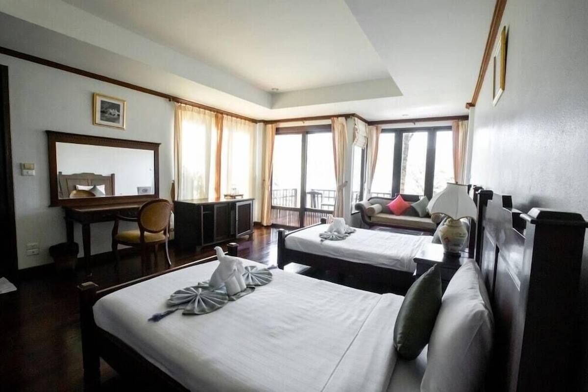 A hotel room with two beds, a desk, a chair, a balcony with curtains, and a large window.