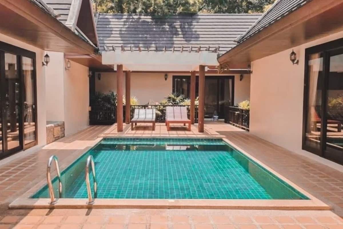 A rectangular swimming pool with a tiled deck, surrounded by a covered patio area with two lounge chairs and a small table.