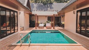 Una piscina rectangular con borde de baldosas, rodeada por un porche cubierto con dos sillones de descanso y una pequeña mesa.